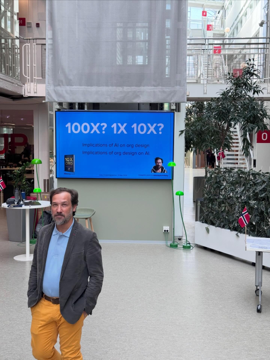 Alexey Krivitsky foran en stand og et banner for boken 10x Org og seminaret Redesign, then AI i Oslo Forskningspark.